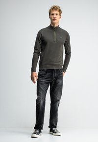 Dunkelgrauer Pullover mit Reißverschluss und gerippten Bündchen, kombiniert mit ausgebleichten schwarzen Jeans und schwarzen Sneakers mit weißen Sohlen.