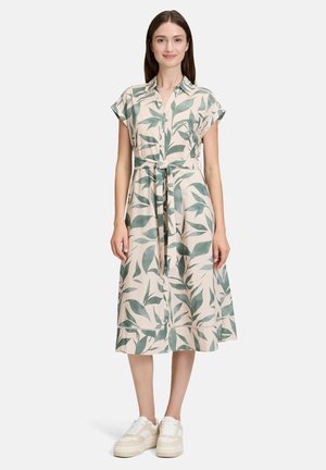 Kwiatowa sukienka w kremowym kolorze z zielonymi wzorami liści, z krótkimi rękawami, zapinana na guziki z przodu, z paskiem do wiązania w talii. Wykończona rozszerzanym dołem.