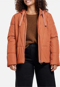 Žena v hnědé péřové bundě, hnědém pleteném svršku a černých kalhotách, usmívá se s kudrnatými vlasy proti bílému pozadí.