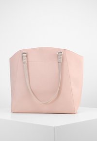 Sac cabas en cuir rose doux avec deux bandoulières rose pâle, présenté sur un piédestal blanc sur un fond uni.