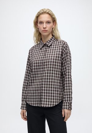 Jeune femme aux cheveux blonds ondulés portant une chemise à carreaux marron et blanc boutonnée et un pantalon noir, debout devant un fond clair uni.