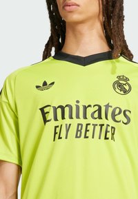 Camiseta de fútbol de color amarillo neón con acentos negros, logo de Adidas y el texto "Emirates FLY BETTER". Tejido texturizado con un diseño de cuello en V.
