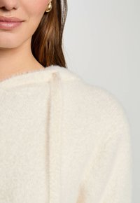 Sudadera color crema hecha de un tejido suave y peludo. Cuenta con un cordón y un diseño simple con una ligera textura.