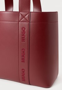 Borsa a spalla in pelle rosso scuro con doppie maniglie e striscia verticale con il logo "HUGO" in rilievo. Texture liscia e costruzione solida.