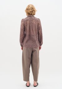 Blusa in pizzo marrone trasparente con motivo floreale, maniche lunghe e colletto classico, abbinata a pantaloni a vita alta color marrone chiaro.