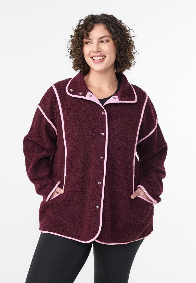 Burgundia fliisjakker, millel on roosa ääre osa, snap-kinnitused ja küljekotid; sel on krae ja vabam lõige lisamugavuse tagamiseks.