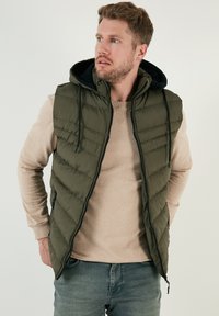 Olivgrön quiltad väst med huva, featuring en blixtlås fram och dragsnören. Bärs över en beige tröja. Avslappnad passform och lätt material.