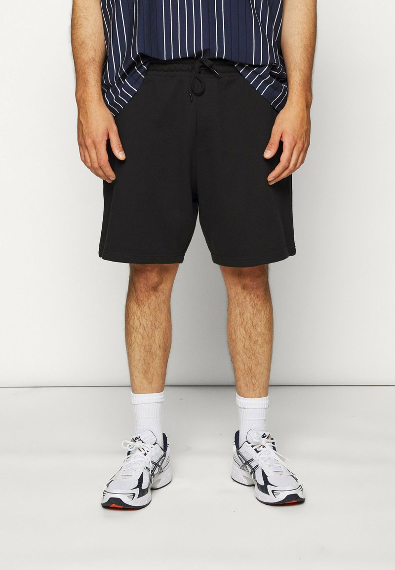 Pantaloni sportivi neri con una cintura regolabile, abbinati a una maglietta a righe blu marino e calze bianche, completati da scarpe da ginnastica.