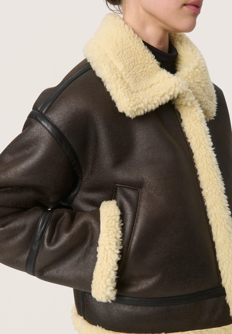 Giacca di pelle marrone con colletto e polsini in pelliccia shearling color crema. Presenta un design strutturato e dettagli accentuati in pelle nera.