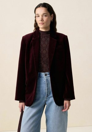 Femme portant un blazer en velours bordeaux foncé sur un haut à motif, assorti à un jean taille haute bleu clair, se tenant devant un fond beige.