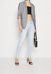Jeans ajustados de color azul claro combinados con un blazer gris de gran tamaño, una camiseta corta negra y tacones negros con tiras. Sosteniendo un pequeño bolso negro.