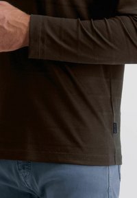 Dunkelbraunes Langarmshirt mit weicher Textur und horizontalen Streifen, mit einem kleinen Markenetikett an der Seite. Dazu trägt es eine blaue Hose.