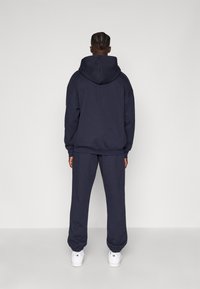 Ναυτικό μπλε φούτερ με κουκούλα και αντίστοιχες φόρμες. Το ύφασμα είναι μαλακό, με ραμμένα μανίκια και μύτες, συνδυασμένο με λευκά αθλητικά παπούτσια.