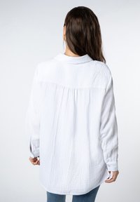 White, textured button-down shirt with a relaxed fit, back pleat, and cuffed long sleeves, worn with light blue jeans.