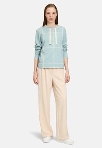 Hellblau gestrickter Pullover mit weißen Nähten und Bindedetail, kombiniert mit beigen weit geschnittenen Hosen und weißen Turnschuhen. Einfaches Design.