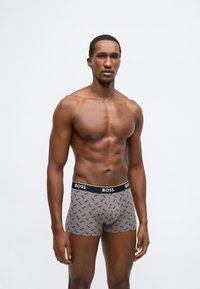 Šedé boxerky s opakujícím se vzorem loga BOSS, elastický černý pas a přiléhavý střih. Model stojí bez trička.