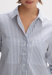 Weißes Hemd mit vertikalen blauen Streifen, klassischem Kragen, Knopfleiste und zwei Brusttaschen. Glatte Baumwolltextur.