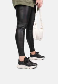 Leggings sintetici neri con finitura lucida; sneakers nere con suole bianche; borsa a tracolla bianca. Le gambe del modello sono visibili dalla metà del polpaccio in giù.