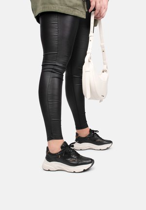Zwarte synthetische leggings met glanzende afwerking; zwarte sneakers met witte zolen; witte schoudertas. De benen van het model zijn zichtbaar van halverwege het daardoor tot beneden.