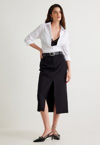 Chemise blanche à boutons portée avec une jupe midi noire avec une fente à l'avant. Ceinture noire avec une boucle ronde. Le mannequin porte des escarpins noirs à bride arrière.