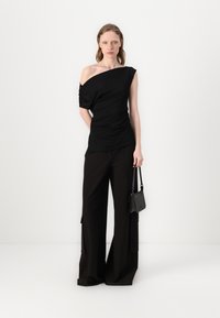 Melns drapēts topiņš ar vienu plecu atstātu brīvu, savienots ar augstās jostas vietas melnām platām biksēm. Neliela melna rokassoma ar plecu siksnu.