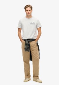 Grå crew-neck t-shirt med grafisk print, beige løse bukser og en mørkternet skjorte bundet om taljen. Lyse sneakers.