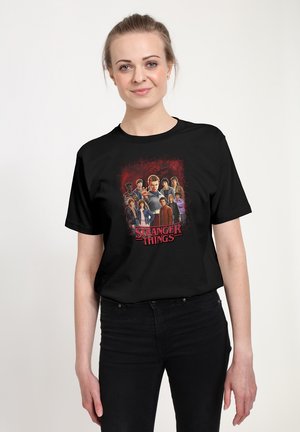Zwart katoenen T-shirt met een kleurrijke afbeelding van personages uit "Stranger Things" tegen een rode achtergrond, gedetailleerd met de titel van de serie.
