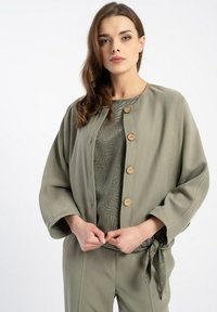 Oliwkowa kurtka z obniżonymi ramionami, dużymi drewnianymi guzikami i wiązanym paskiem. Pod spodem, teksturowany zielony top.