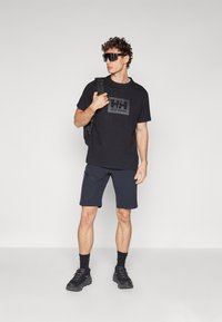 Tricou negru Helly Hansen cu un logo pe piept, asortat cu pantaloni scurți negri și adidași negri, purtând un rucsac.