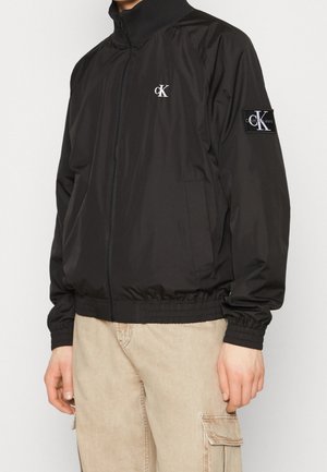 Osoba nosząca czarną kurtkę zapinaną na zamek Calvin Klein z logo na piersi i rękawie, w połączeniu z beżowymi spodniami typu cargo.