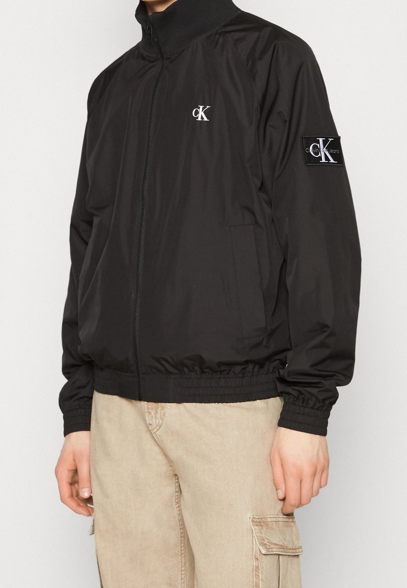 Personne portant une veste zippée noire Calvin Klein avec un logo sur la poitrine et la manche, associée à un pantalon cargo beige.