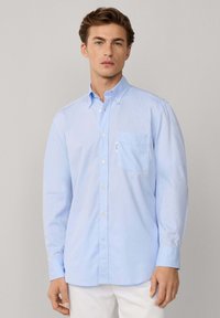 Camisa de algodón azul claro de botones, con un bolsillo en el pecho, cuello clásico y mangas largas con puños abotonados. Textura suave.