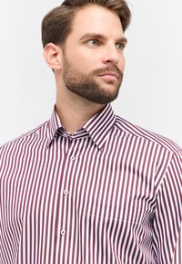 Uomo con barba marrone curata che indossa una camicia bianca con strisce verticali color bordeaux, che guarda leggermente verso destra su uno sfondo semplice.