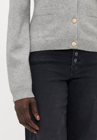 Gray knit cardigan with ribbed cuffs, featuring two front pockets and three decorative buttons, paired with black denim jeans.