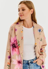 Béžový kvetinový blazer s ružovými, fialovými a oranžovými kvetmi; štruktúrovaný dizajn s jemnou látkou, nosený cez biely top a džínsy.