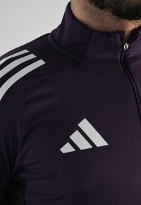 Sudadera deportiva morada con cremallera, fabricada en una tela suave, que presenta un diseño de tres rayas blancas en el hombro y un logotipo en el pecho.