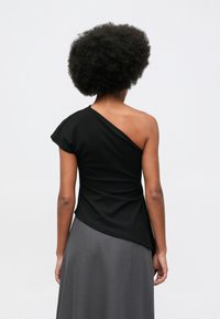 Kobieta z fryzurą afro, ubrana w czarną bluzkę na jedno ramię i szary spódnicę, zwrócona plecami w stronę jasnego, jednolitego tła.