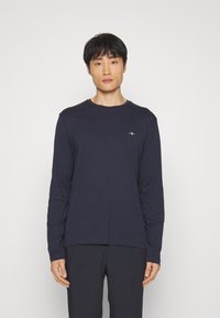 T-shirt à manches longues bleu marine en tissu doux, avec un col rond et un petit logo sur la poitrine. Porté avec un pantalon foncé.