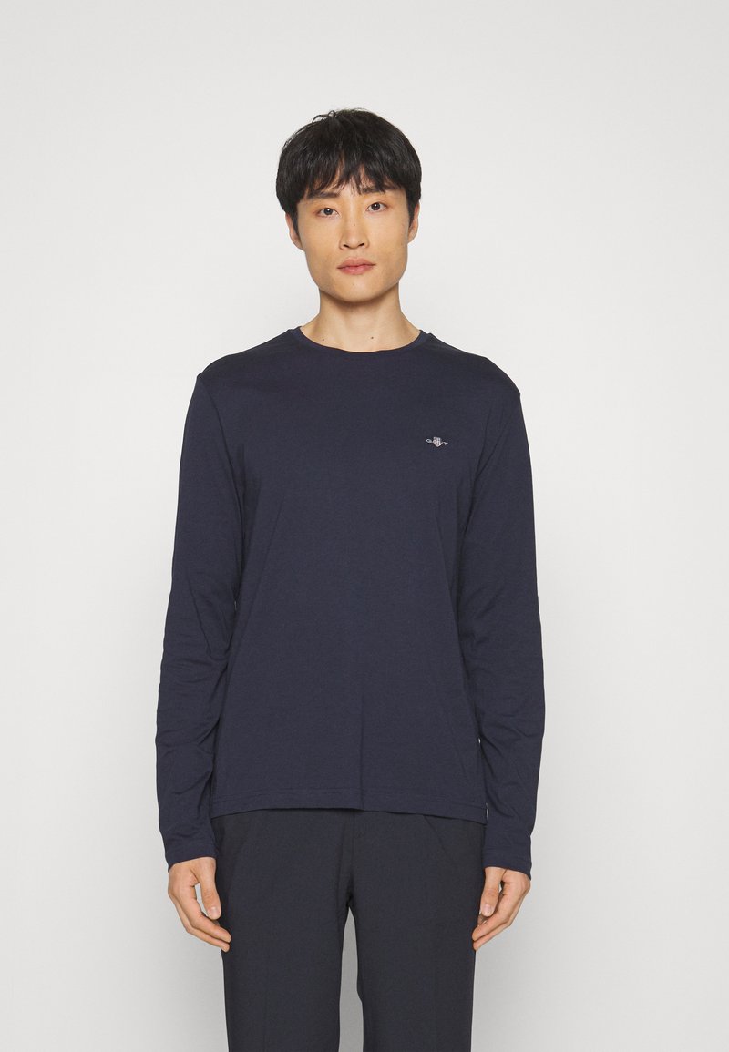 T-shirt à manches longues bleu marine en tissu doux, avec un col rond et un petit logo sur la poitrine. Porté avec un pantalon foncé.