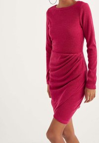 Abito aderente in maglia rosa con maniche lunghe, scollo rotondo e dettagli drappeggiati in vita. Texture liscia e lunghezza a metà coscia.