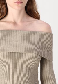 Beige aversettet ribbestrikket topp med lange ermer, med en bred krage og teksturert strikkemønster. Lite logo detalj nær kanten.