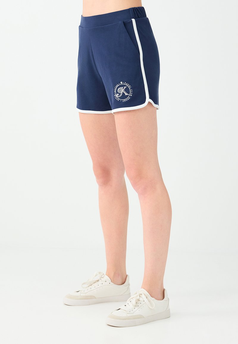 Personne portant un short de sport bleu marine avec une bordure blanche et des baskets blanches, debout devant un fond clair uni.