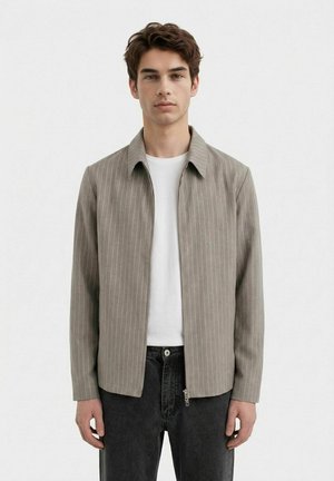 Ung mann iført en lys brun stripete glidelåsjakke over en hvit t-skjorte og svarte jeans, stående mot en ensfarget bakgrunn.