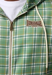 Green plaid zip-up hoodie with white, yellow, and dark green lines. Features a logo patch and a cream-colored zipper with a gold pull.