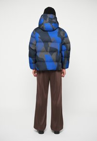 Jachetă puffer camuflaj albă și gri cu glugă, având un design matlasat și textură netedă, combinată cu pantaloni lejeri maro cu talie înaltă.