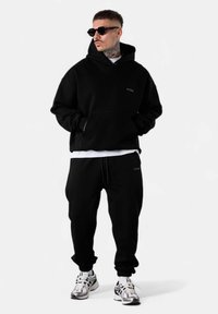Schwarzer Hoodie und passende Sweatpants aus weichem Material, mit einer Fronttasche und minimalem Branding. Kombiniert mit reflektierenden Sneakers.