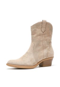 Bota de cowboy de ante beige con punta afilada, costuras decorativas y pequeños detalles de tachuelas. Cuenta con un tacón bajo apilado y lengüetas para facilitar el calzado.