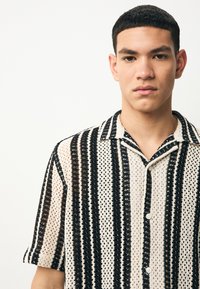 Camisa de manga corta con un patrón de rayas negras y crema, hecha de un material tipo malla con botones al frente y detalles en el cuello.