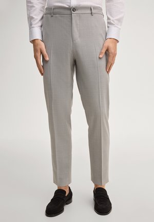 Uomo che indossa pantaloni su misura grigio chiaro e mocassini neri in suede, in piedi con le mani vicino ai fianchi contro uno sfondo semplice.