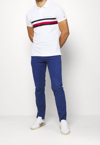 Hvid polo skjorte med en marineblå og rød vandret stribe, marineblå bukser og hvide sneakers med røde og blå detaljer.